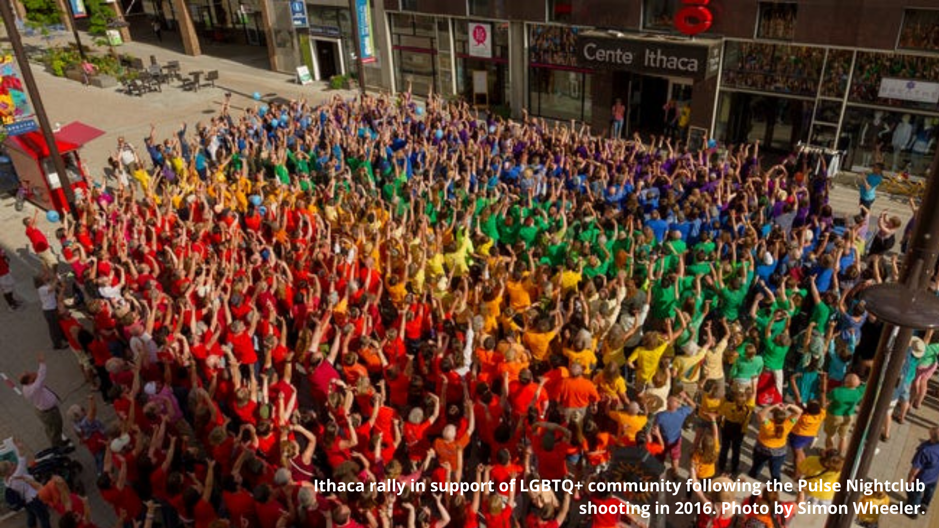 An image of a large crowd wearing red, orange, yellow, green, blue and purple shirts to make a rainbow. The caption says "Ithaca rally support of LGBTQ+ community following the Pulse Nightclub shooting in 2018. Photo by Simon Wheeler."