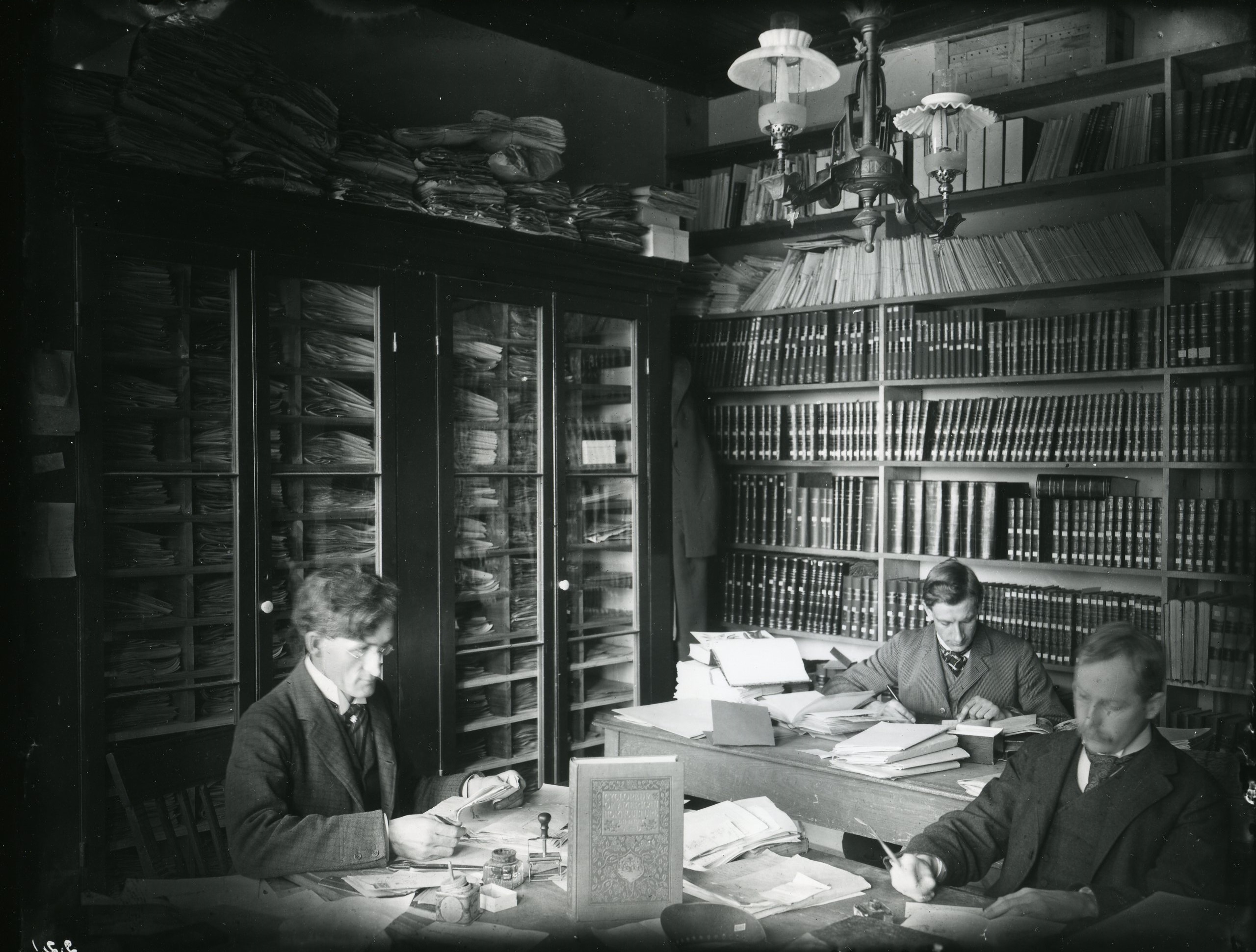 A rectangular black and white image of men at tables surrounded by books.