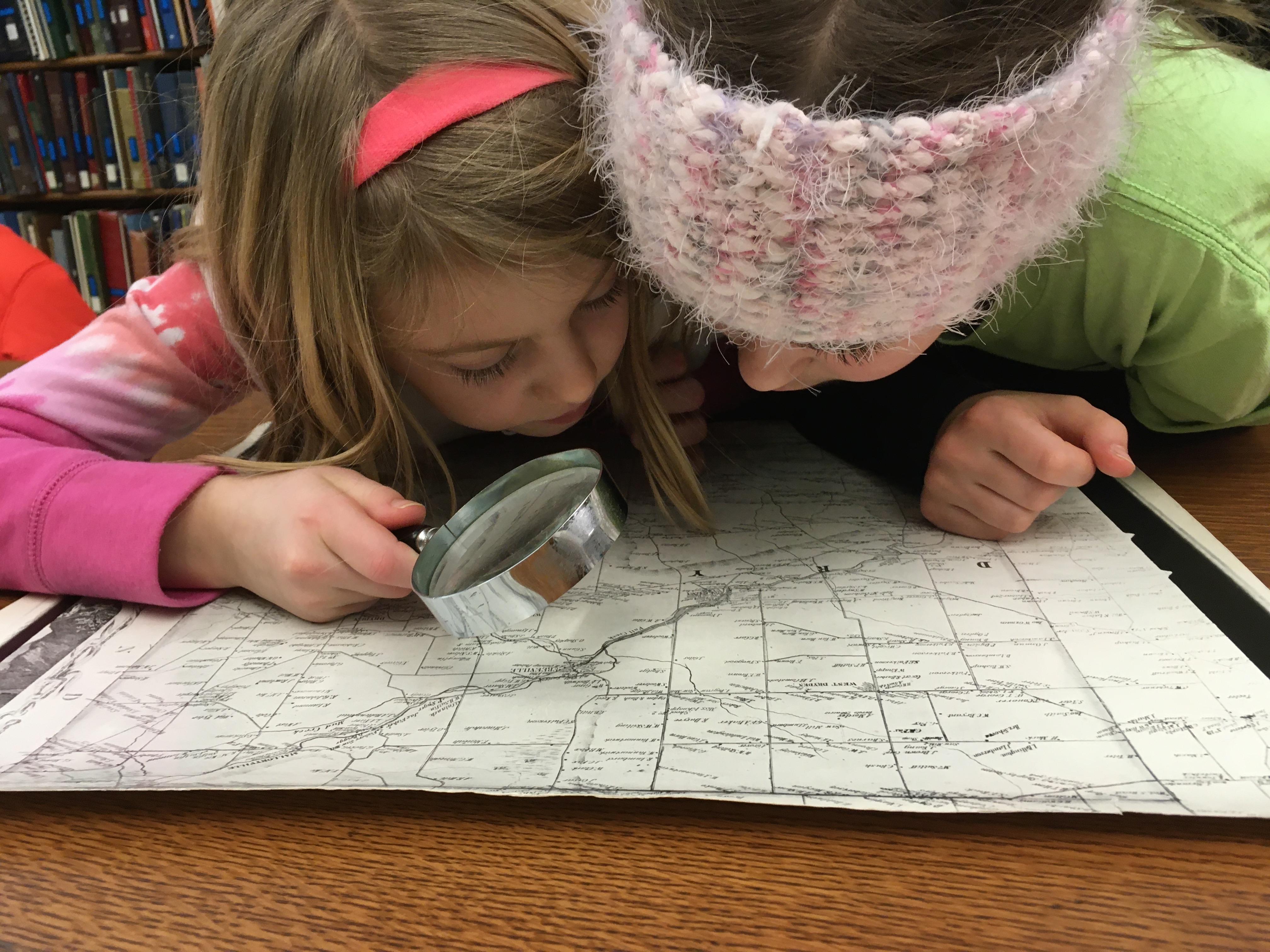An image of two young girls looking at a map. One girl holds a magnifying glass.