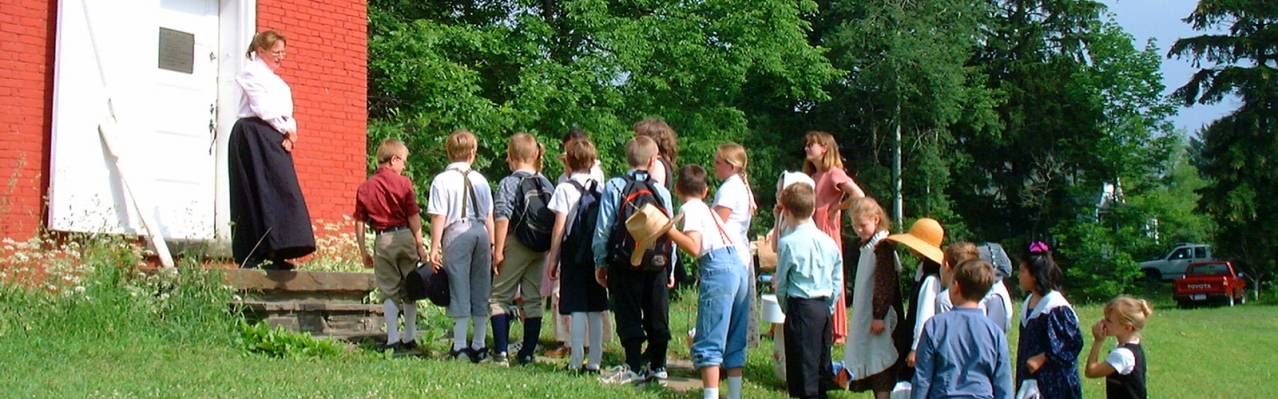 A photo of a group of young children in a line outside of the Eight Square Schoolhouse, accompanied by a teacher.